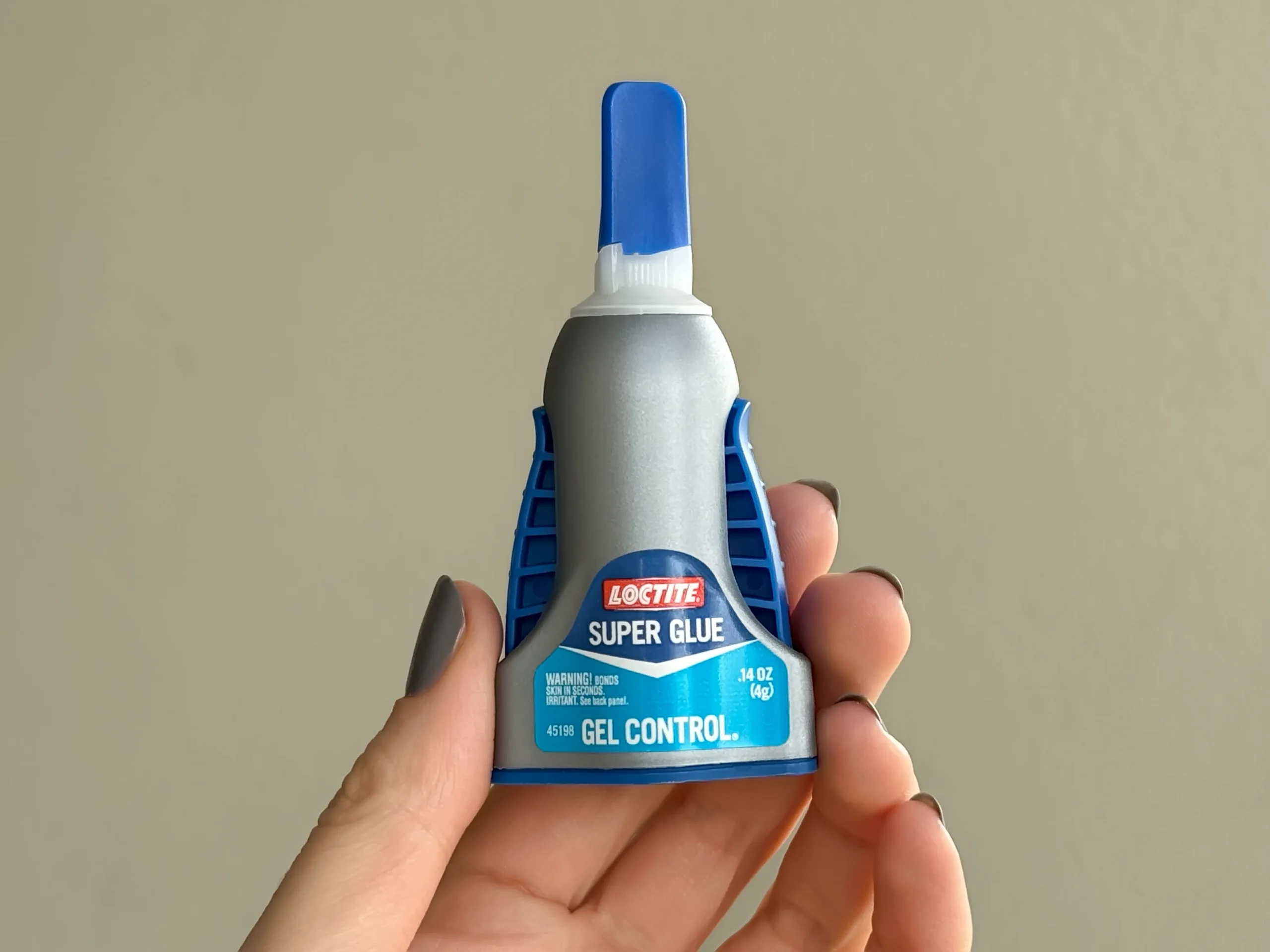 A hand holding a bottle of Loctite Super glue for polymer clay. The bottle is gray, with light blue label. The cap is darker blue. On each side of the bottle, there are dark blue buttons that dispense glue when pushed on.