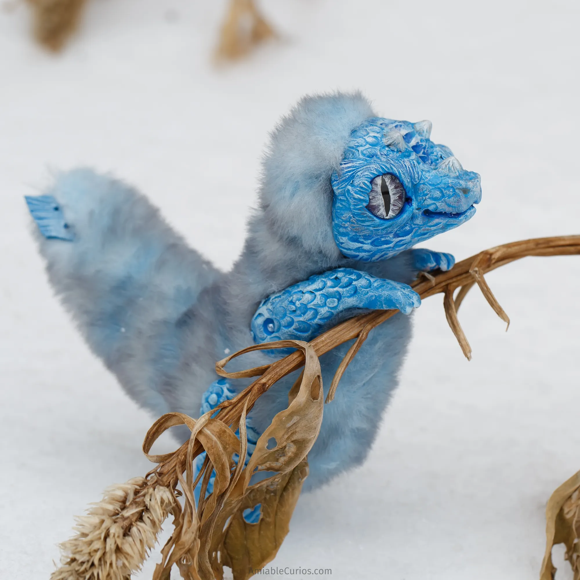 Photo of a handmade unique blue dragon art doll. The dragon is standing on a snowy landscape on his back paws. His front paws are on a dry plant, he's looking over it. 